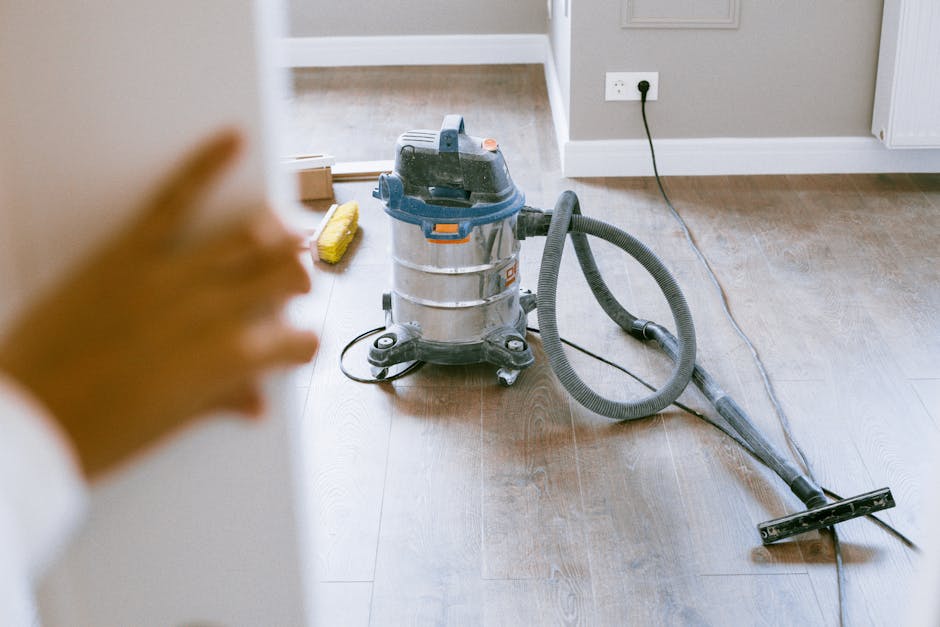 A close-up view of a commercial vacuum cleaner with a metal and plastic body, sitting on a wooden floor during a surface cleaning process, with a person’s hand in the foreground. The vacuum hose is extended across the floor, and a cleaning attachment is visible. The background shows an electrical socket with a plug inserted and a dustpan on the floor. The room features neutral-colored walls and baseboards, and the floor surface appears clean and well-maintained, indicative of professional domestic cleaning services by Cleaner Merton as part of their apartment cleaning guide for Mitcham Broadway, CR4, Merton.