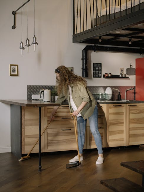A woman with long curly hair dressed in casual clothing, including a beige jacket, white top, and blue jeans, is vacuuming a hardwood floor in a modern kitchen. The kitchen features wooden cabinets with a natural finish, a stainless steel sink, and a countertop with a kettle and some small appliances. The room is well-lit with natural and ambient lighting, including three pendant lights hanging from the ceiling. There are framed pictures on the white wall, and a lofted area with a black railing above. Cleaner Merton’s professional cleaning services focus on surface cleaning and maintaining hygiene in residential spaces like this, ensuring thorough deep cleaning and sanitisation.
