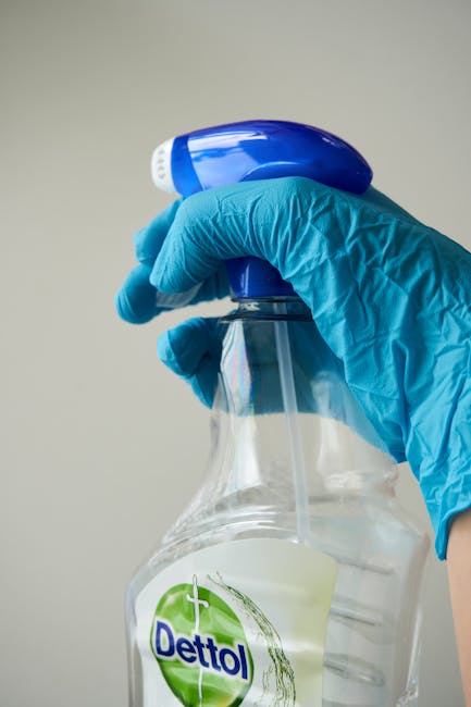 Close-up of a person cleaning a smooth, white surface with a white cloth, wearing blue nitrile gloves. The individual holds a spray bottle, branded 'LooClear', containing a cleaning solution, aimed at disinfecting the area. The lighting is bright, emphasizing the cleanliness and shine of the surface, which appears free of dust and stains. The background is plain, focusing attention on the detailed cleaning process, illustrating hygiene and surface sanitation in line with domestic cleaning and deep cleaning practices, as promoted by Cleaner Merton on their website.
