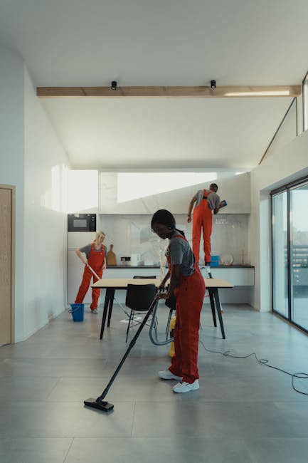Interior view of a modern, minimally decorated kitchen during a professional cleaning session, featuring light-colored ceramic floor tiles and white walls with large floor-to-ceiling windows that allow natural light to flood the space. Three cleaners, wearing red overalls, are engaged in surface cleaning activities: one using a vacuum cleaner on the tiled floor, another wiping the kitchen counter, and a third cleaning the high window or ceiling area. The kitchen includes a black dining table with chairs, a white backsplash, and sleek cabinetry, showcasing a clean and well-maintained environment. The scene emphasizes thorough domestic cleaning by Cleaner Merton, highlighting comprehensive surface cleaning, dust removal, and sanitisation processes in a residential setting.