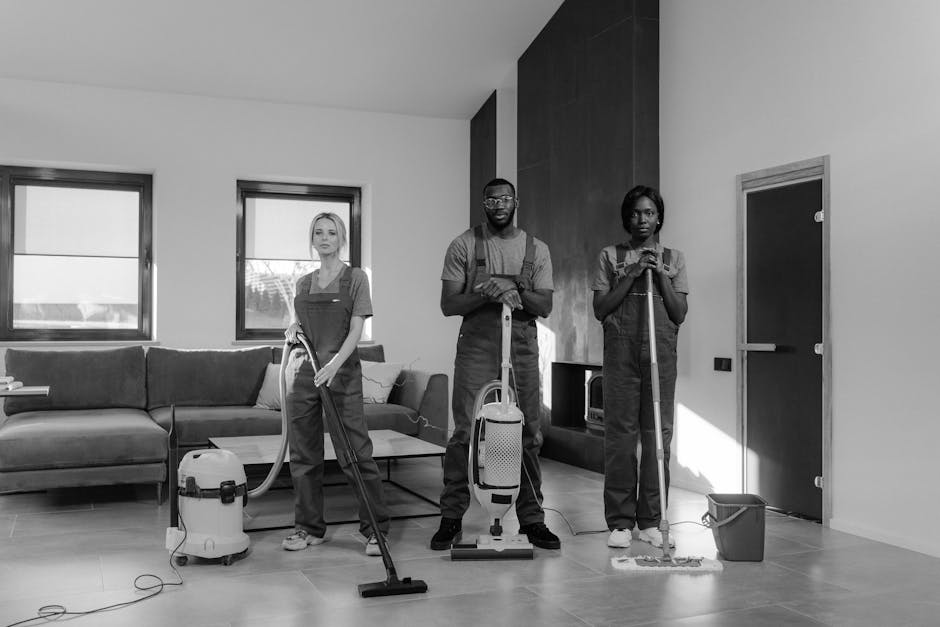 Interior view of a modern, minimally decorated kitchen during a professional cleaning session, featuring light-colored ceramic floor tiles and white walls with large floor-to-ceiling windows that allow natural light to flood the space. Three cleaners, wearing red overalls, are engaged in surface cleaning activities: one using a vacuum cleaner on the tiled floor, another wiping the kitchen counter, and a third cleaning the high window or ceiling area. The kitchen includes a black dining table with chairs, a white backsplash, and sleek cabinetry, showcasing a clean and well-maintained environment. The scene emphasizes thorough domestic cleaning by Cleaner Merton, highlighting comprehensive surface cleaning, dust removal, and sanitisation processes in a residential setting.