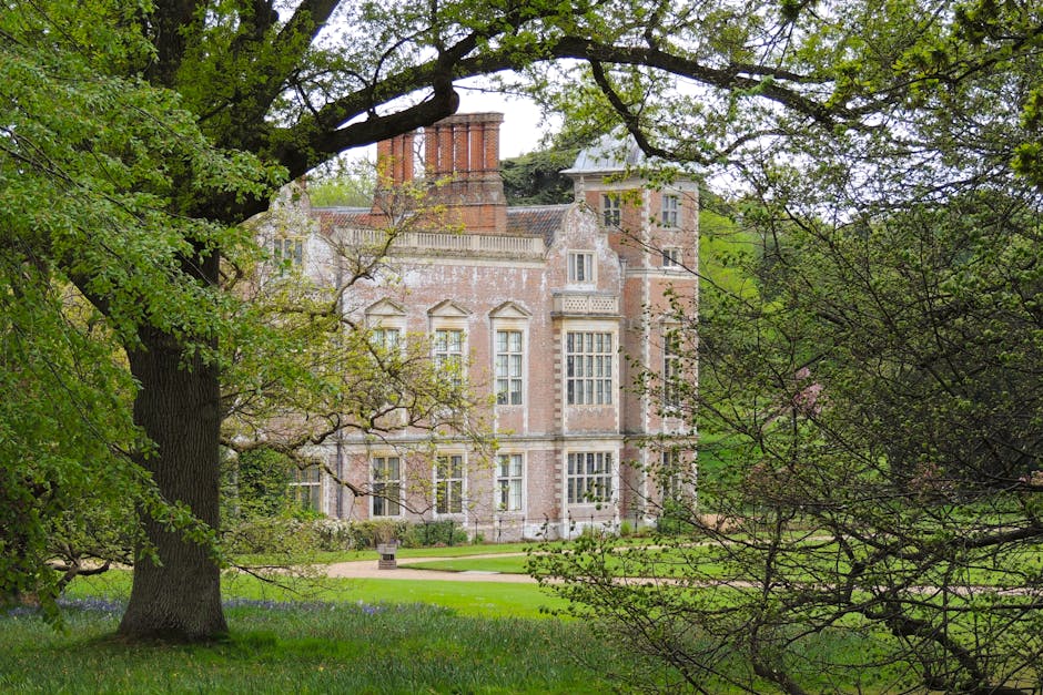 A large, historic stone mansion situated on a well-maintained lawn with pathways, surrounded by mature trees with bare branches, under a partly cloudy sky. The building features multiple chimneys, tall rectangular windows, and a symmetrical facade, indicative of traditional architectural style. In the foreground, the grassy area appears to be lightly frosted or damp, with a clear walking path leading toward the entrance. The scene is illuminated by soft natural light, creating a calm and pristine atmosphere, reflecting the importance of cleanliness and maintenance in residential settings. As part of cleaning practices for homes on Morden Hall Park Estate, tasks may include surface cleaning of exterior stonework, window washing, and turf maintenance, which [COMPANY_NAME] provides to ensure the property's aesthetic and hygiene standards are upheld. This image emphasizes the significance of thorough exterior and grounds cleaning as part of maintaining a beautiful, hygienic home environment, aligned to the themes of surface cleaning, deep cleaning, and sanitisation discussed on the webpage.
