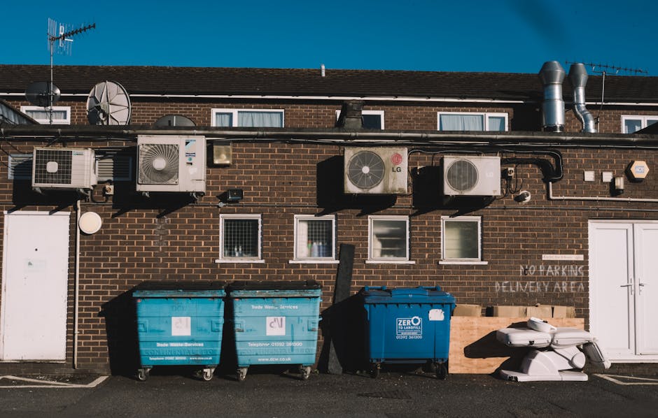 Exterior view of a brick building with multiple air conditioning units and ventilation pipes installed on the upper wall. The building features four small rectangular windows with white frames and a white door on the left side. In front, three large wheelie bins—two blue and one darker blue—are positioned on the pavement, with a pile of white discarded furniture or equipment placed beside them. The scene is lit with natural daylight, highlighting the cleanliness and orderliness of the area. The image reflects a well-maintained commercial waste disposal site, as seen in the context of disposal of commercial waste in Merton, with the company Cleaner Merton providing cleaning and waste management services.