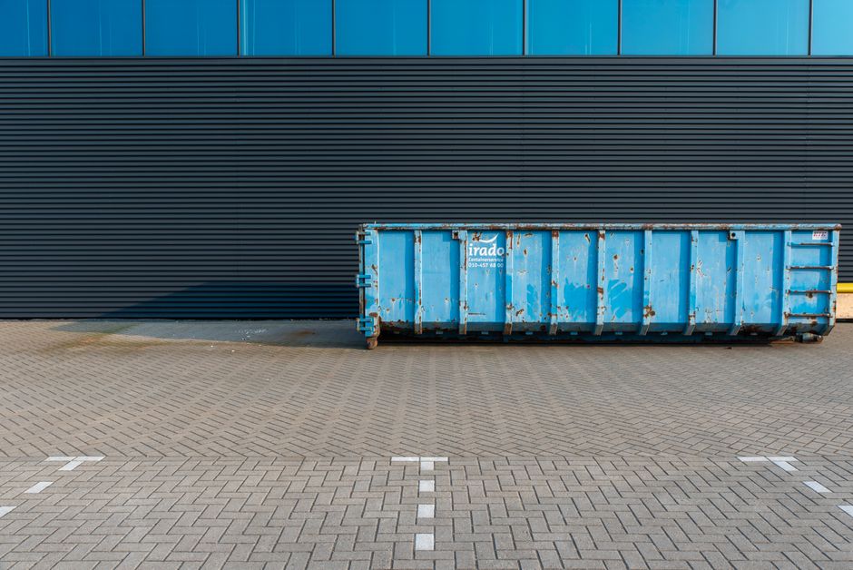 A weathered blue commercial waste container with rust patches and chipped paint sits on a paved area outside a modern building with blue and black horizontal paneling. The container is positioned near the building wall, casting a shadow on the ground, which is made of interlocking concrete pavers. The area appears clean and well-maintained, with no visible debris around. The lighting suggests a sunny day, illuminating the scene clearly. This image exemplifies waste management infrastructure often associated with commercial cleaning and disposal services offered by Cleaner Merton, aligned with the page about disposing of commercial waste in Merton, including permit and fines information.