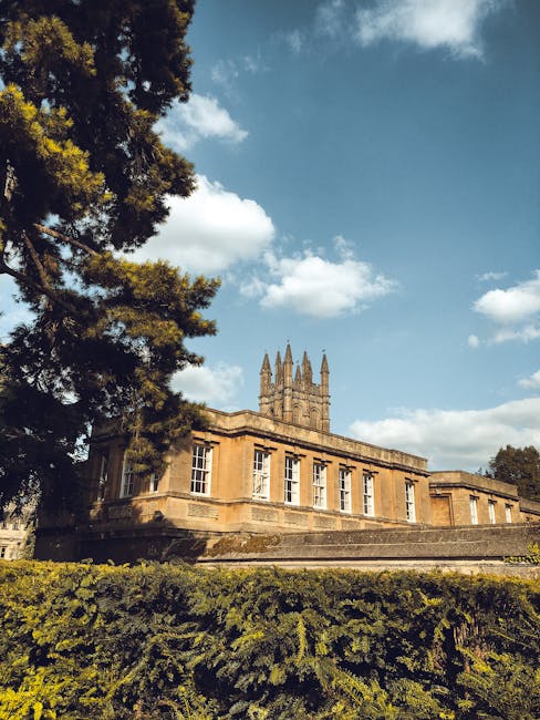A historic stone building with large windows and a prominent tower featuring multiple spires, set against a partly cloudy sky with scattered white clouds. The structure is partially obscured by a tall tree on the left and a well-maintained hedge in the foreground, indicating its location within a landscaped area. The building's exterior appears clean and well-preserved, with sunlight highlighting its architectural details, suggesting regular maintenance and scenic surroundings. This setting relates to the importance of cleanliness and upkeep, aligning with domestic and commercial cleaning themes as per Merton Council's rules for household waste and cleaning, as promoted by Cleaner Merton.