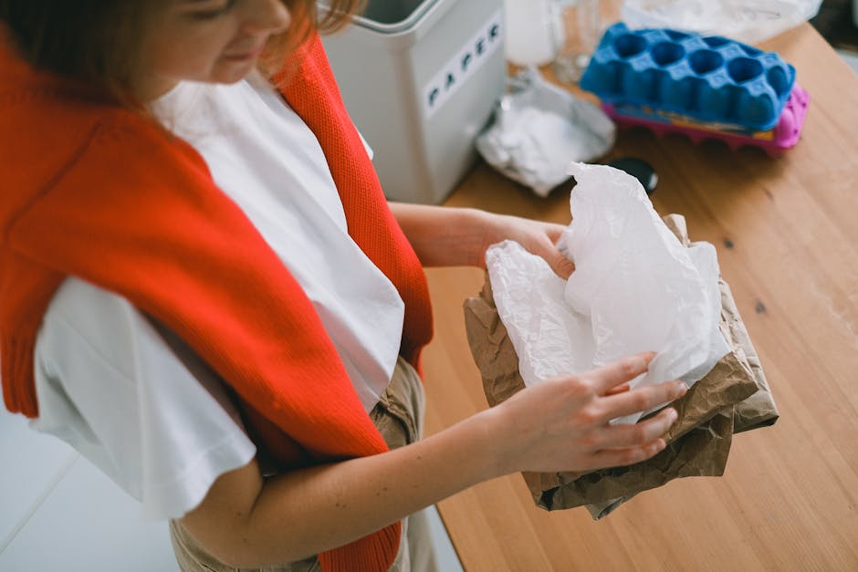 A person wearing a white shirt and a red knitted vest is holding a brown paper bag with a white paper tissue or wiping cloth inside, positioned over a wooden table. The table surface is clean and has a natural wood finish. In the background, there is a blue ice-cube tray, a white appliance, and a roll of white paper towel, indicating a setting related to domestic cleaning. The lighting is natural, highlighting the tidy and hygienic environment. This image reflects surface cleaning and hygiene practices, consistent with the themes of household waste management and cleaning guidelines detailed in Merton Council rules for household waste and cleaning, as provided by Cleaner Merton.