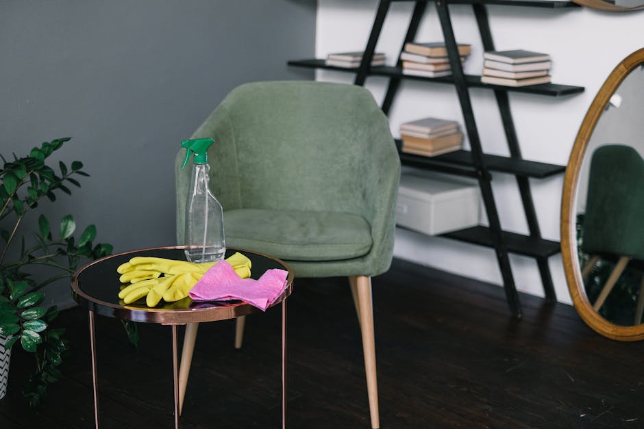 A person wearing light green gloves and a khaki jacket is cleaning a dark wooden bookshelf in a room with soft, diffuse lighting. They are reaching up to dust or arrange a row of hardcover books, some