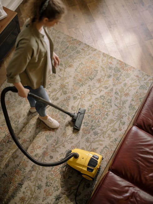 A woman using a yellow vacuum cleaner to deep clean a patterned area rug in a living room with wooden flooring and a red leather sofa. She is dressed casually in a beige jacket, jeans, and white shoes, focusing on surface cleaning and removing dust from the carpet. The room is well-lit, highlighting the cleanliness of the rug and surrounding floor, demonstrating professional domestic cleaning services by Cleaner Merton near Colliers Wood High Street.