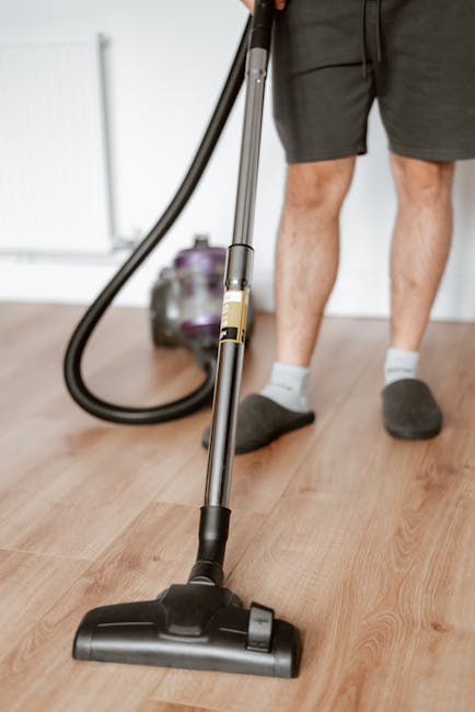 A woman using a yellow vacuum cleaner to deep clean a patterned area rug in a living room with wooden flooring and a red leather sofa. She is dressed casually in a beige jacket, jeans, and white shoes, focusing on surface cleaning and removing dust from the carpet. The room is well-lit, highlighting the cleanliness of the rug and surrounding floor, demonstrating professional domestic cleaning services by Cleaner Merton near Colliers Wood High Street.