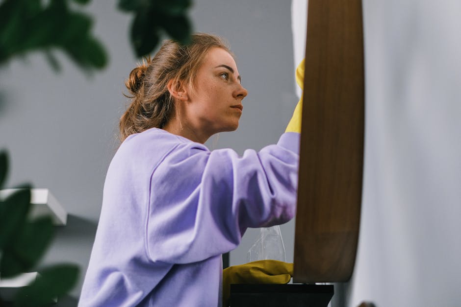 A woman with light brown hair tied back, wearing a lavender sweatshirt and yellow rubber gloves, is focused on cleaning a wooden surface with a cloth in a well-lit room. The surface appears to be a ta
