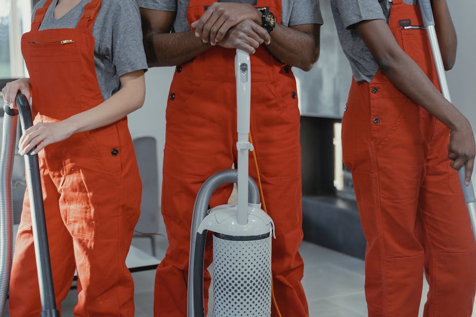 Close-up of three professional cleaners dressed in grey shirts and bright orange overalls, holding vacuum cleaners with grey hoses and white and grey canister units. The image focuses on their midsections, showing their hands gripping the vacuum handles and their matching uniforms. They are standing indoors on a tiled floor, with a modern background including a window and furniture. The scene highlights surface cleaning and sanitisation processes, emphasizing the importance of deep cleaning in residential or commercial spaces. The presence of cleaning tools and the neat, dust-free environment aligns with the services offered by Cleaner Merton, as promoted on their website at cleanermerton.co.uk, under a page discussing the costs and benefits of professional cleaning services.